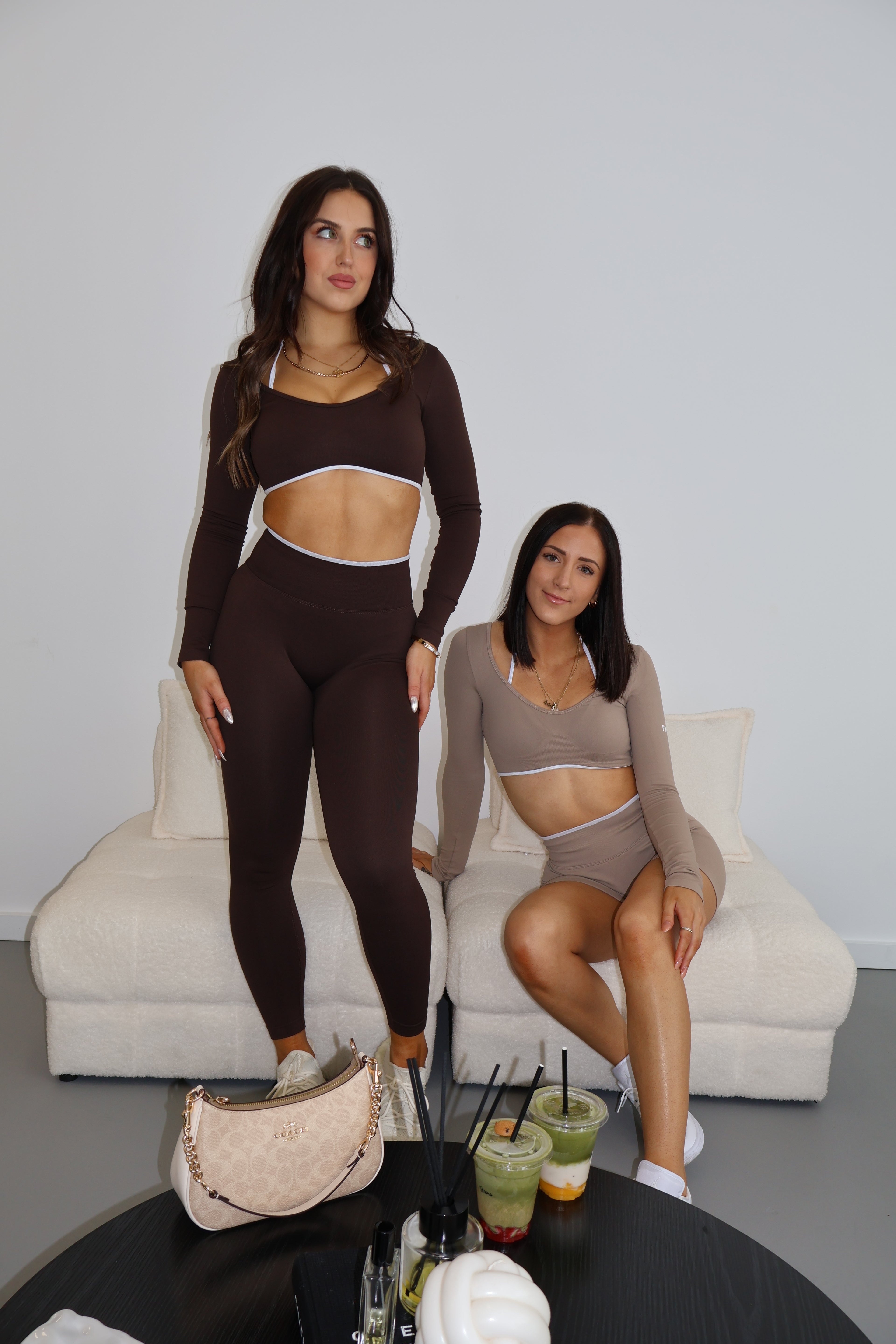 Two women in matching activewear sets standing on a white couch against a gray wall. Brown activewear sets. Viral scrunch shorts.